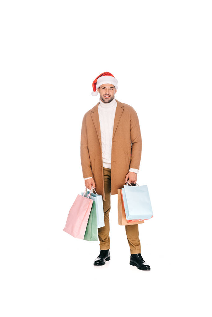 bello barbuto giovane uomo in cappello di Babbo Natale tenendo borse della spesa e sorridendo alla fotocamera isolata su bianco
 - Foto, immagini