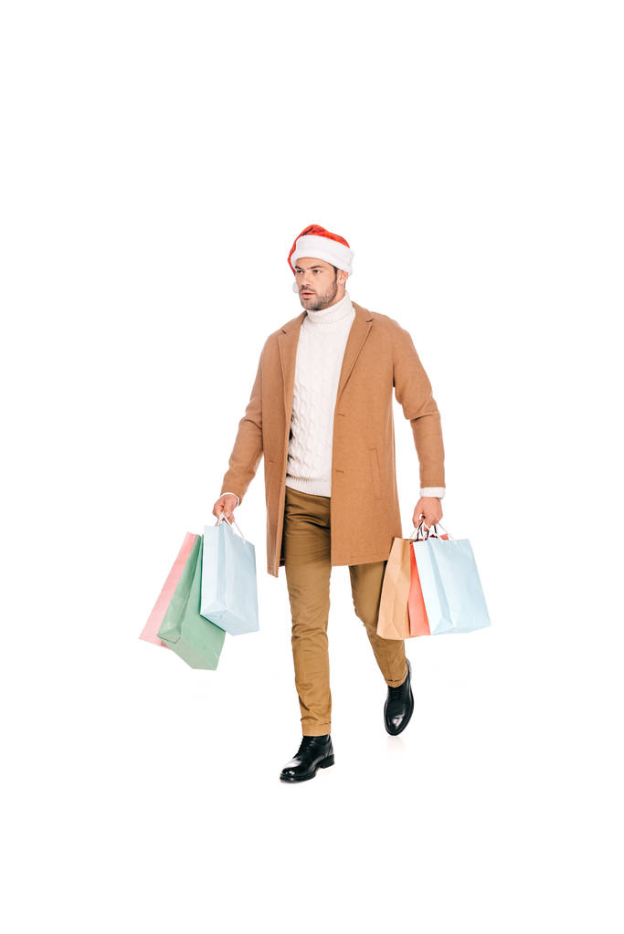Widok pełnej długości młody człowiek w santa hat przytrzymanie torby na zakupy i spacer na białym tle - Zdjęcie, obraz