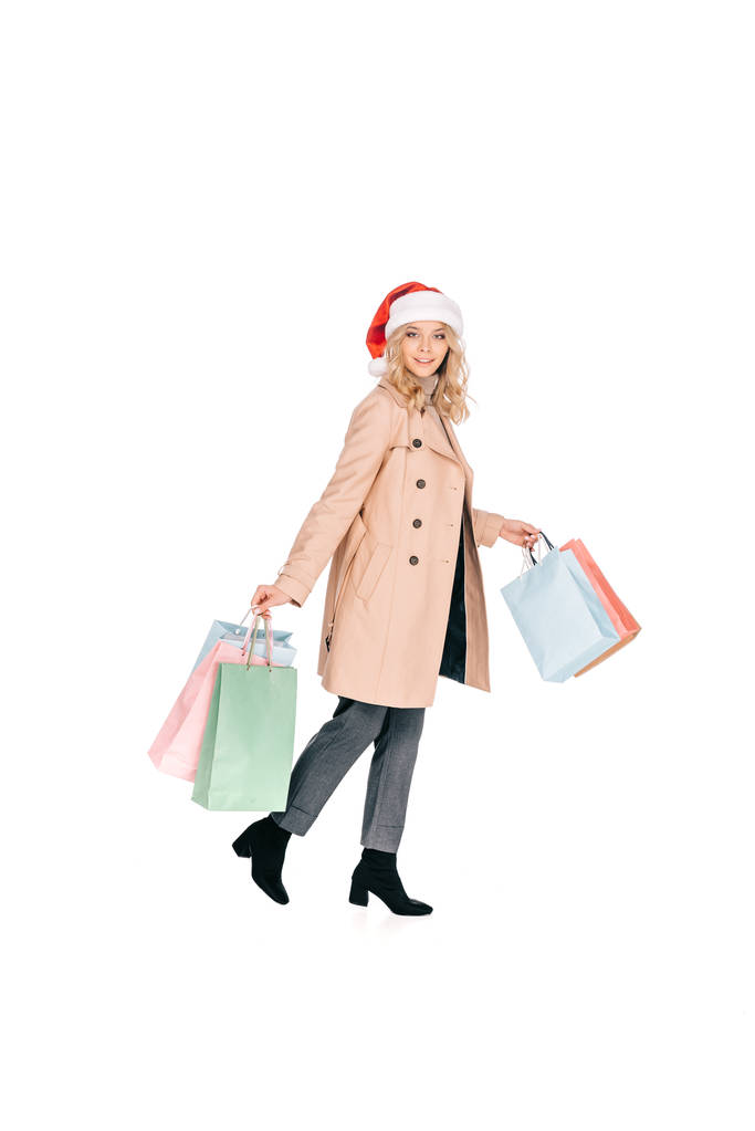 full length view of woman in santa hat walking with shopping bags and smiling at camera isolated on white - Photo, Image