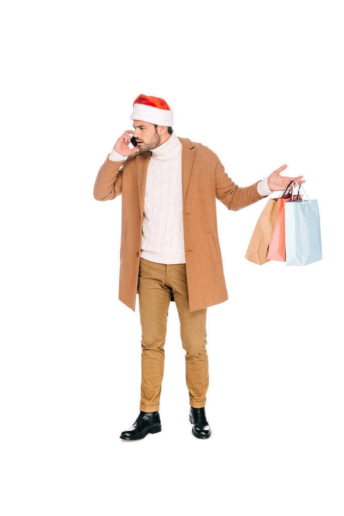 эмоциональный молодой человек в шляпе Санты держит сумки с покупками и разговаривает по смартфону изолированы на белом
 - Фото, изображение