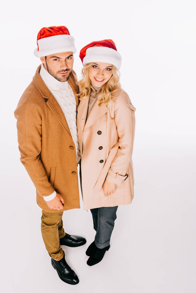 Hochwinkelaufnahme des schönen jungen Paares in Weihnachtsmannmützen, die in die Kamera lächeln, isoliert auf weiß - Foto, Bild