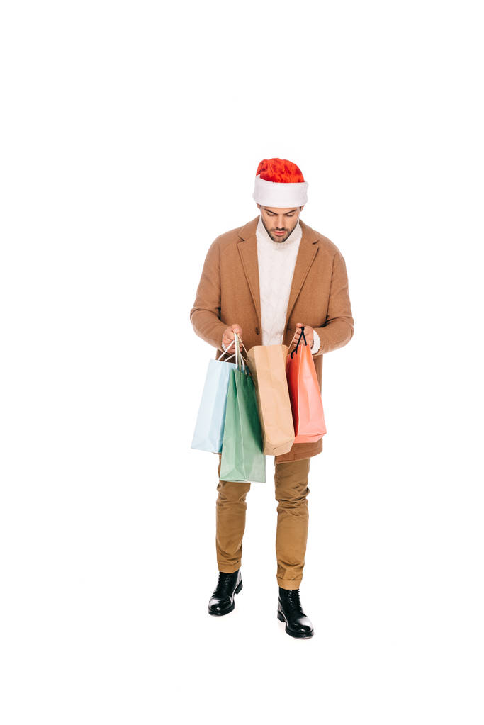 Молодой человек в шляпе санта в полный рост держит в руках сумки с покупками, изолированные на белом фоне
 - Фото, изображение