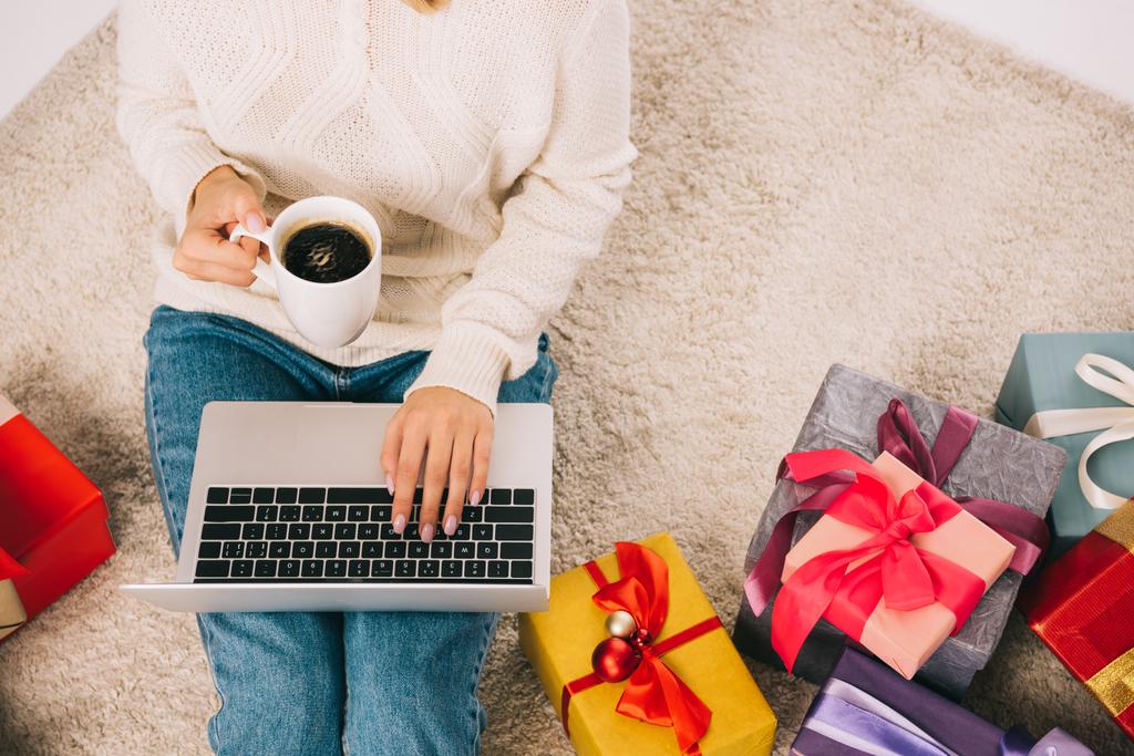 vue partielle du dessus de la jeune femme tenant une tasse de café et utilisant un ordinateur portable tout en étant assis près des cadeaux de Noël
 - Photo, image