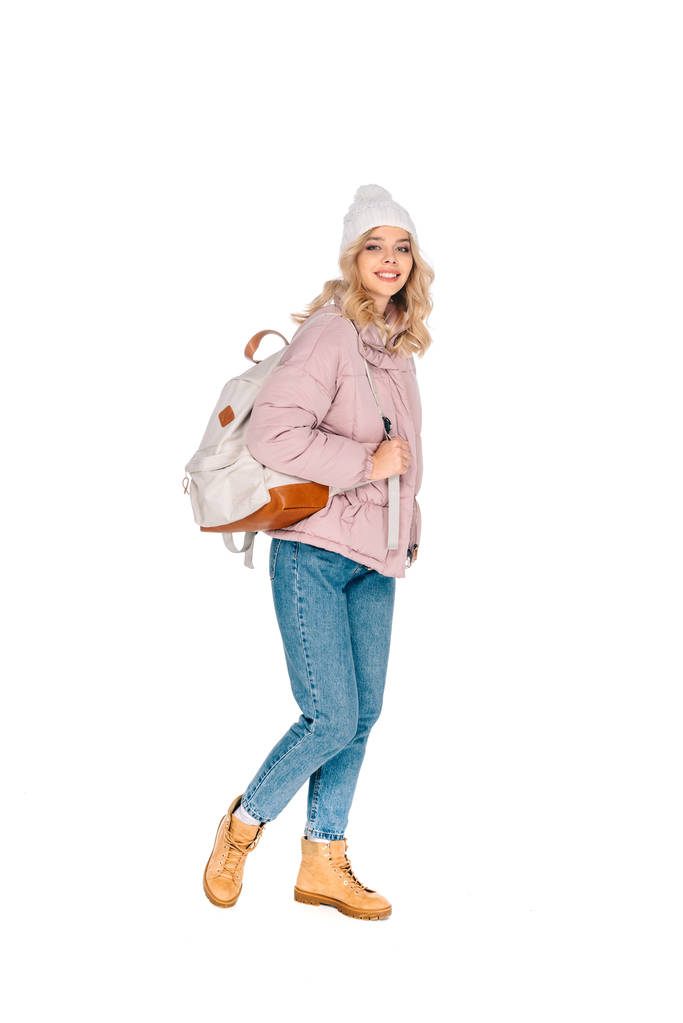 όμορφη νεαρή γυναίκα ταξιδιώτης στο καπέλο περπάτημα με σακίδιο και να χαμογελά στη φωτογραφική μηχανή που απομονώνονται σε λευκό - Φωτογραφία, εικόνα