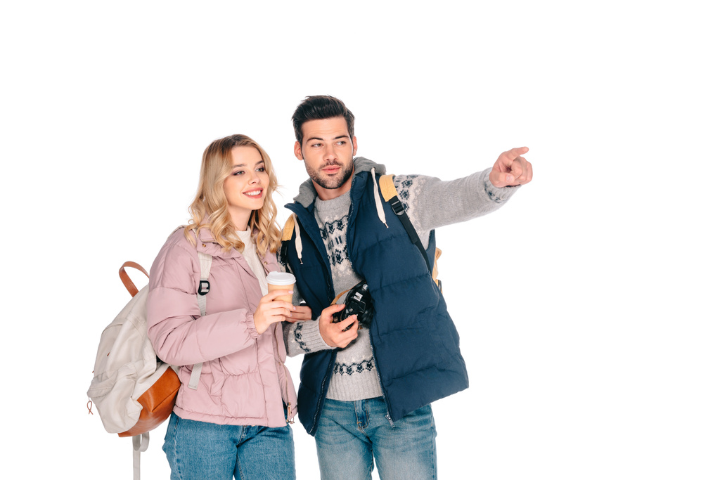 joven pareja de turistas con mochilas sosteniendo café para ir y la cámara mientras señala y mira hacia otro lado aislado en blanco
 - Foto, Imagen