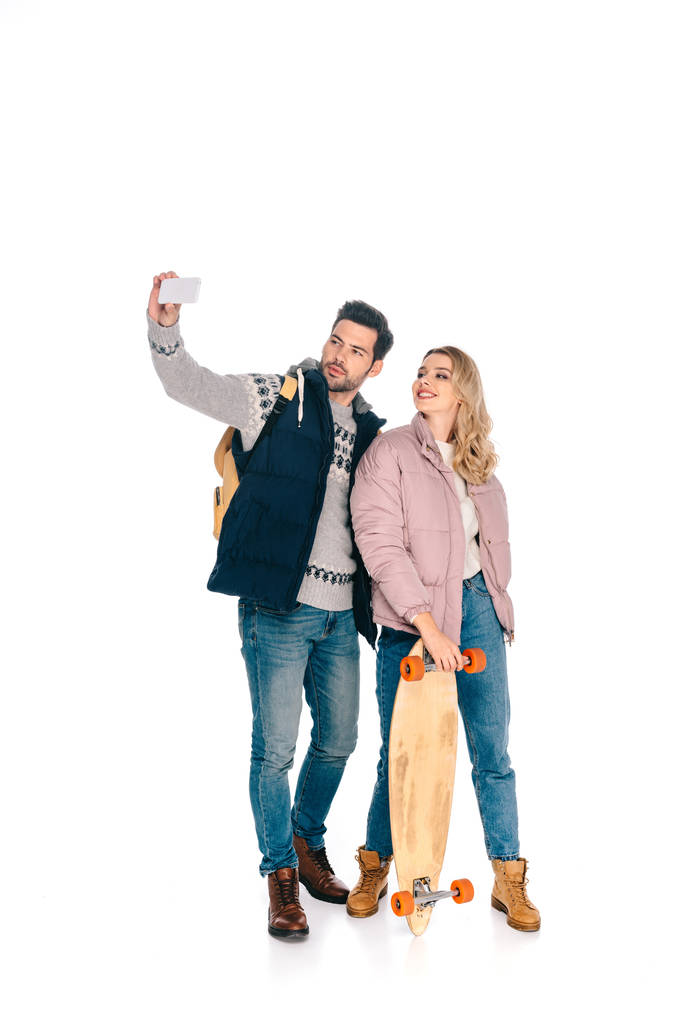 smiling young couple with backpack and skateboard taking selfie with smartphone isolated on white - Photo, Image