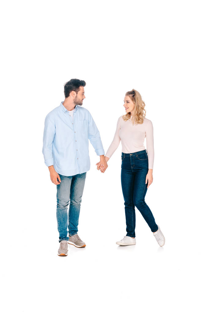 Вид в полный рост счастливой молодой пары, держащейся за руки и улыбающейся друг другу на белом фоне
 - Фото, изображение