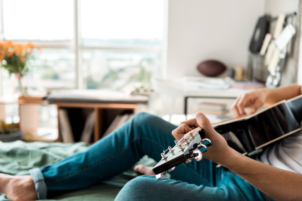 Schnappschuss von Gitarrist in Jeans, der zu Hause auf dem Bett Akustikgitarre spielt - Foto, Bild