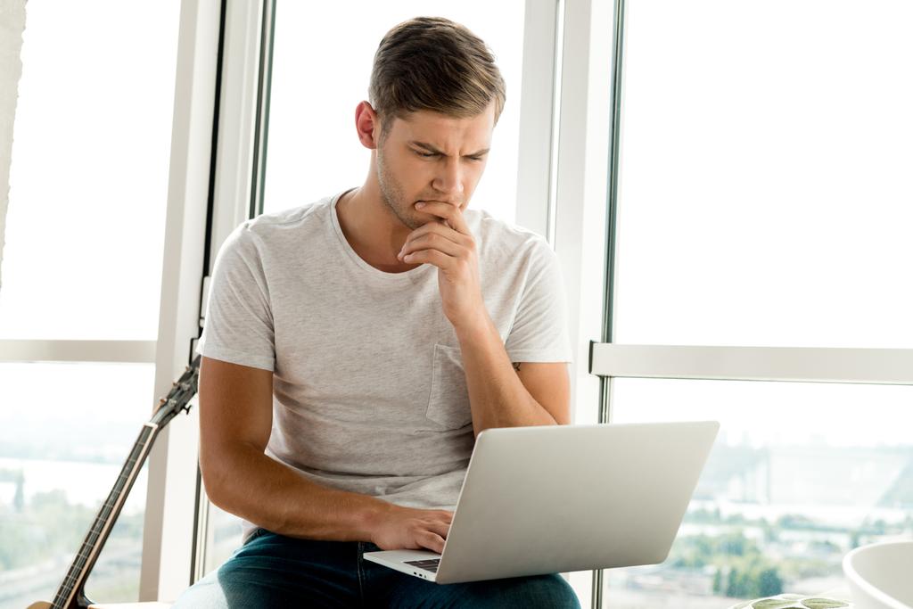 зосереджений чоловік у повсякденному одязі, використовуючи ноутбук, сидячи у вікні вдома
 - Фото, зображення