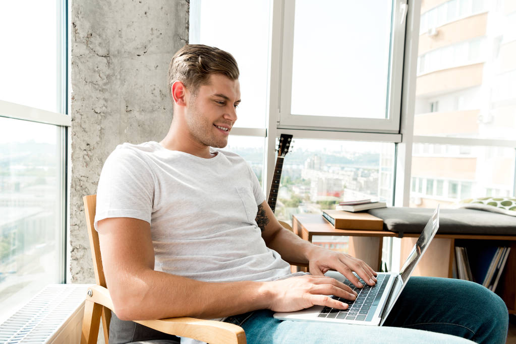 Glimlachende man in leunstoel laptop thuis gebruiken - Foto, afbeelding