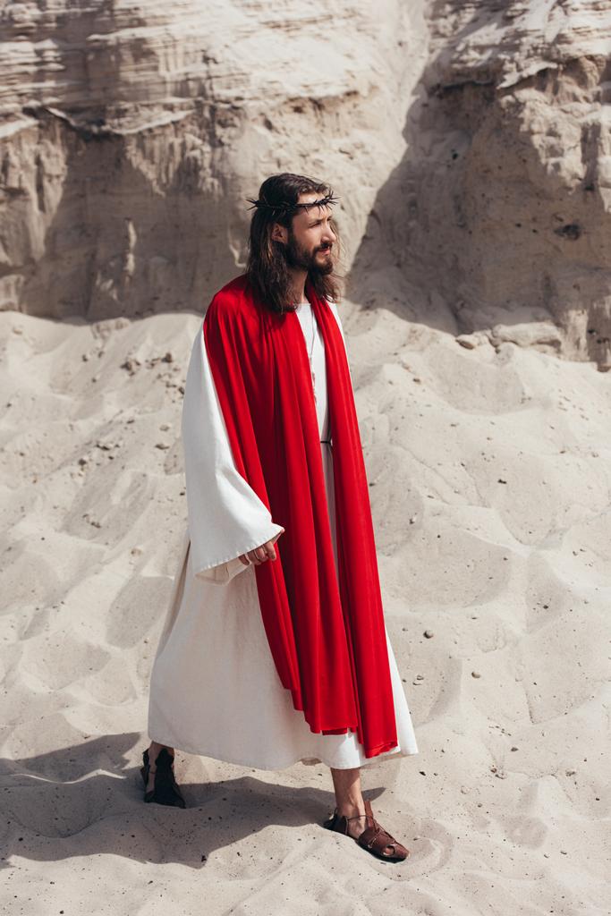 vista lateral de Jesús en túnica, faja roja y corona de espinas caminando en el desierto
 - Foto, imagen