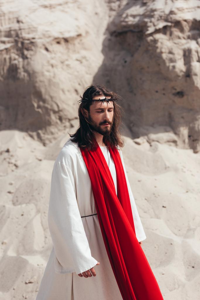 bonito Jesus em roupão, faixa vermelha e coroa de espinhos andando com os olhos fechados no deserto
 - Foto, Imagem