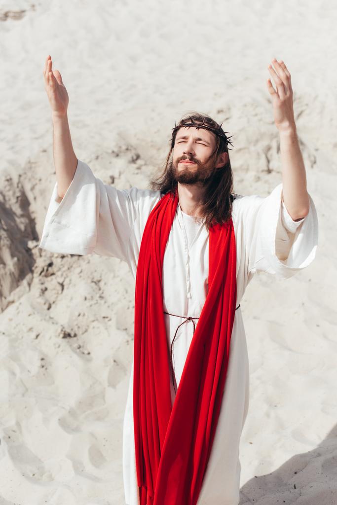 Jesus in robe, red sash and crown of thorns standing with raised hands and praying in desert - Photo, Image
