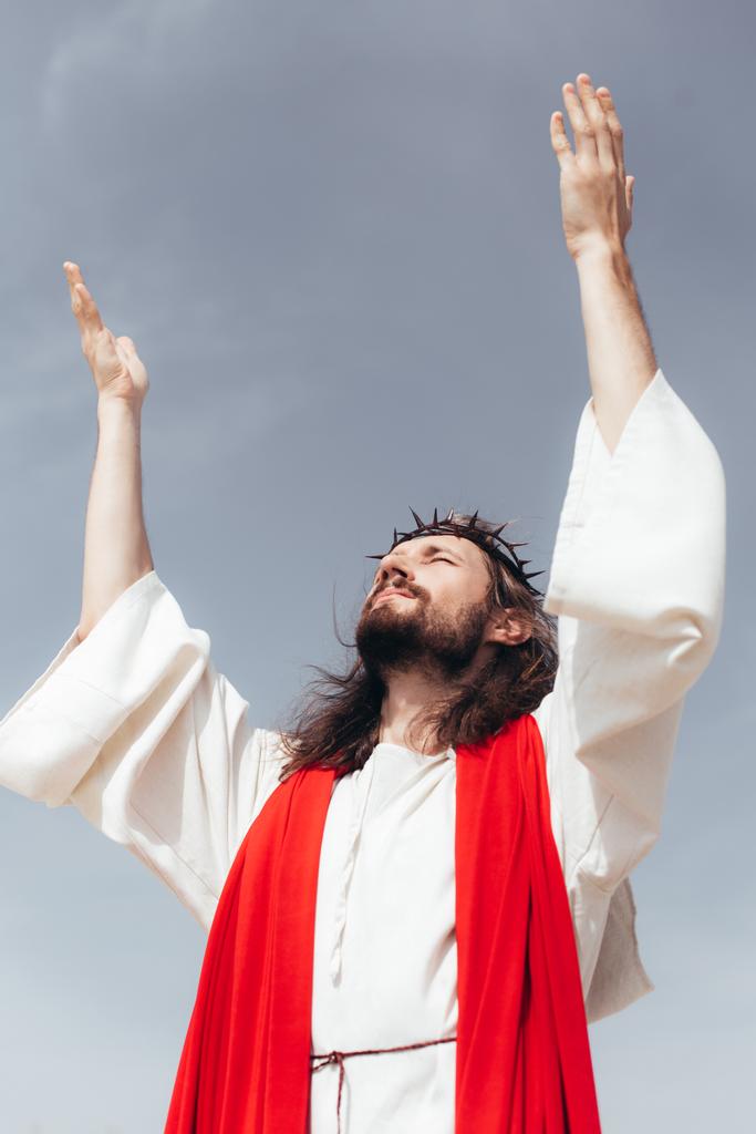 vista angolo basso di Gesù in veste, fascia rossa e corona di spine in piedi con le mani alzate contro il cielo grigio
 - Foto, immagini