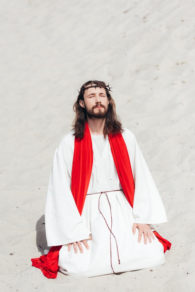 Jesus in Robe, roter Schärpe und Dornenkrone steht auf Knien mit geschlossenen Augen und betet in der Wüste - Foto, Bild
