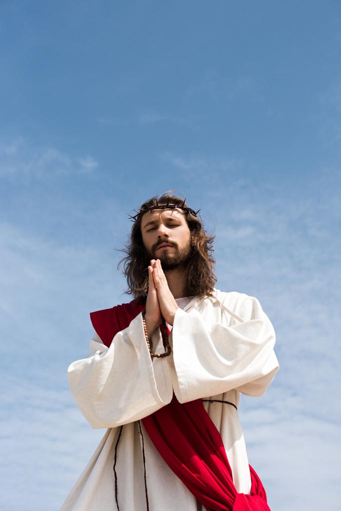 vista de ángulo bajo de Jesús en túnica, faja roja y corona de espinas sosteniendo rosario y orando con los ojos cerrados contra el cielo azul
 - Foto, imagen