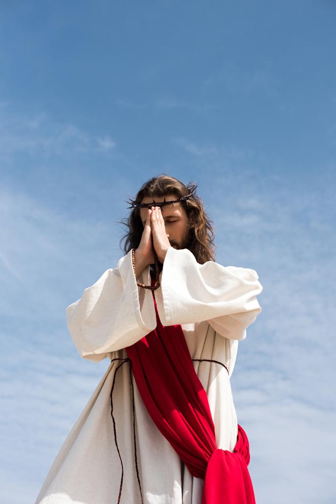 vista de ángulo bajo de Jesucristo en túnica, faja roja y corona de espinas sosteniendo rosario y orando contra el cielo azul
 - Foto, imagen