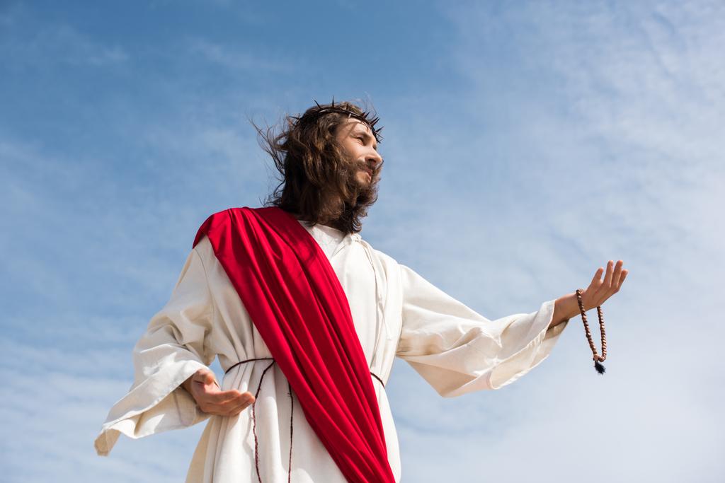 nízký úhel pohled na Ježíše v šat drží růženec a dosažení ruku proti modré obloze - Fotografie, Obrázek