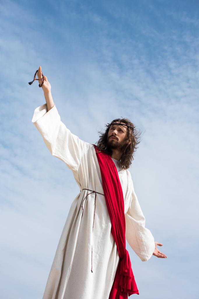 nízký úhel pohled na Ježíše v roucho, červenou šerpou a trnová koruna drží růženec a stál s otevřenou náručí proti modré obloze - Fotografie, Obrázek