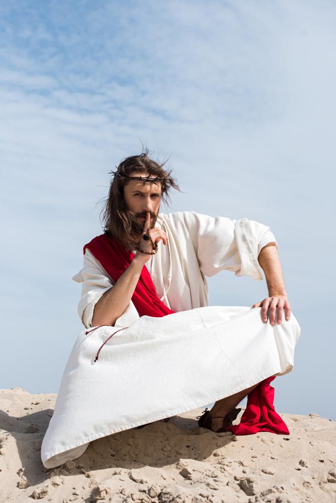 Jesus em roupão, faixa vermelha e coroa de espinhos segurando rosário e mostrando sinal de silêncio no deserto
 - Foto, Imagem