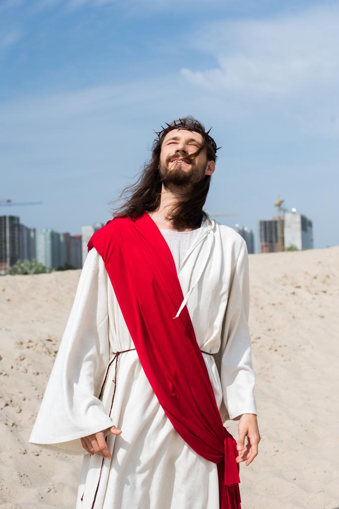 Иисус в мантии, красной ленте и терновом венце стоял на песке с закрытыми глазами, здания на заднем плане
 - Фото, изображение