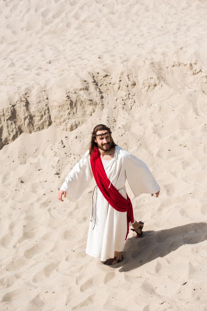 high angle view of Jesus in robe, red sash and crown of thorns walking on sand in desert - Photo, Image