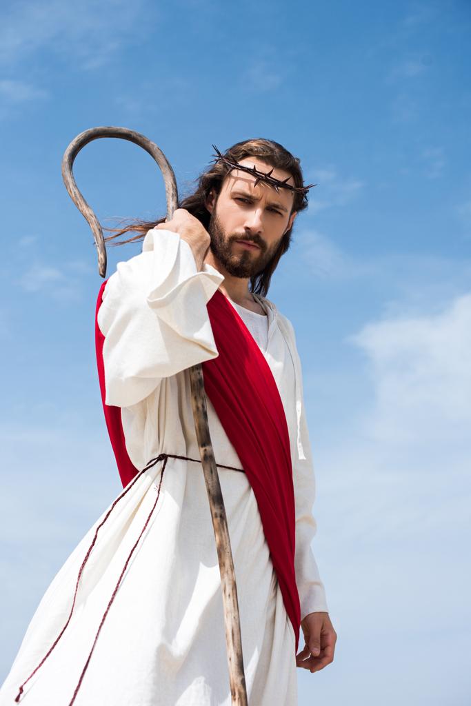 vue en angle bas de Jésus en robe, ceinture rouge et couronne d'épines debout avec le bâton en bois dans le désert et regardant la caméra
 - Photo, image
