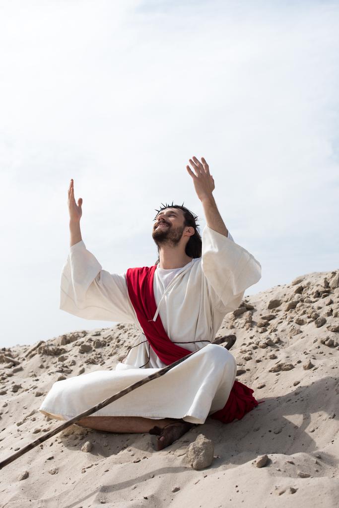 smiling Jesus in robe, red sash and crown of thorns sitting in lotus position with raised hands and talking with god on sand in desert - Photo, Image