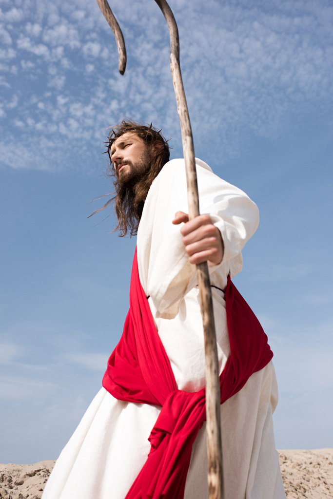vista de ángulo bajo de Jesús en túnica, faja roja y corona de espinas caminando en el desierto con bastón
 - Foto, Imagen