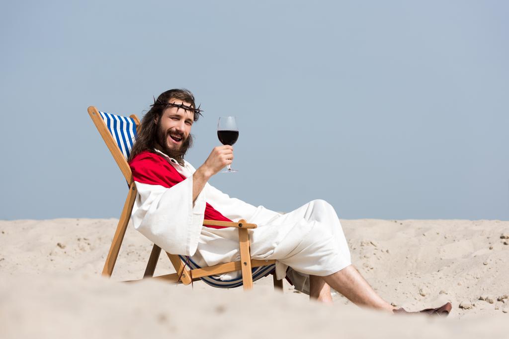mosolygós Jézus köntös és pihenő nyugágyon pohár vörös bor, a sivatagban, látszó-on fényképezőgép piros káva - Fotó, kép