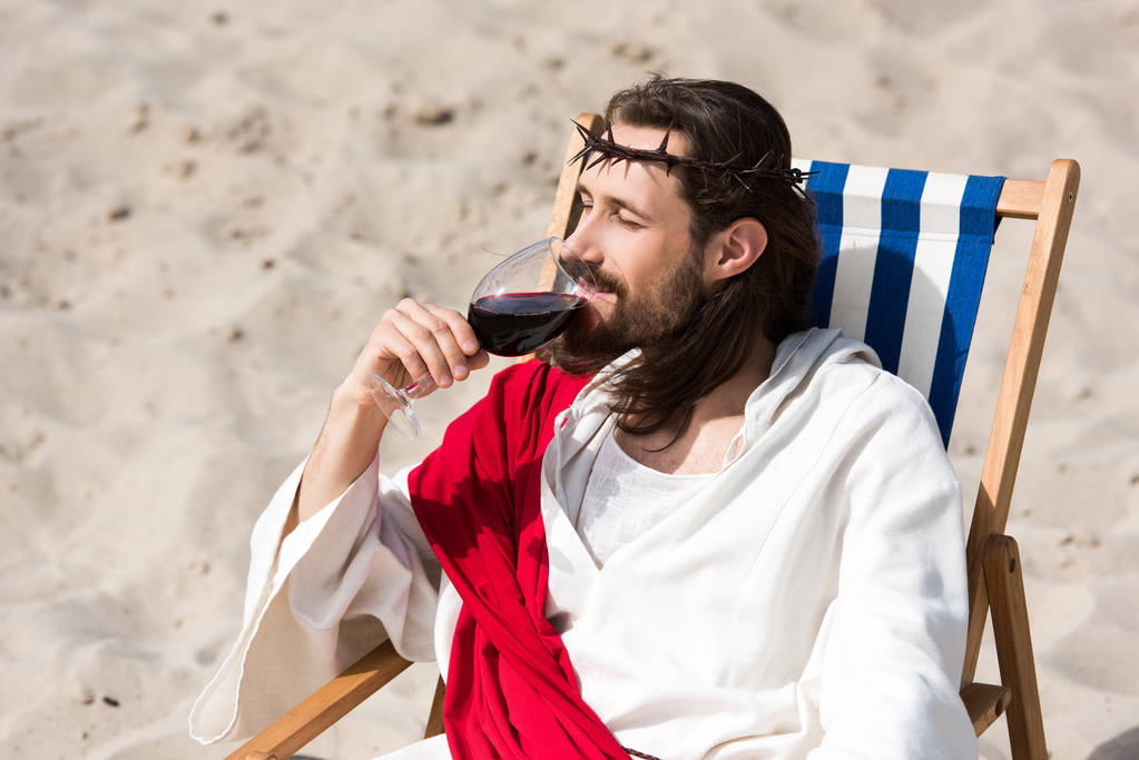 Ο Ιησούς ρόμπα και κόκκινο φύλλο αναπαύεται σε ξαπλώστρα και πίνοντας κόκκινο κρασί στην έρημο - Φωτογραφία, εικόνα