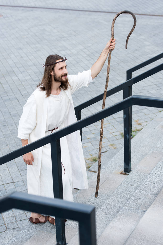 high angle view of cheerful Jesus in robe and crown of thorns walking on stairs with staff - Photo, Image