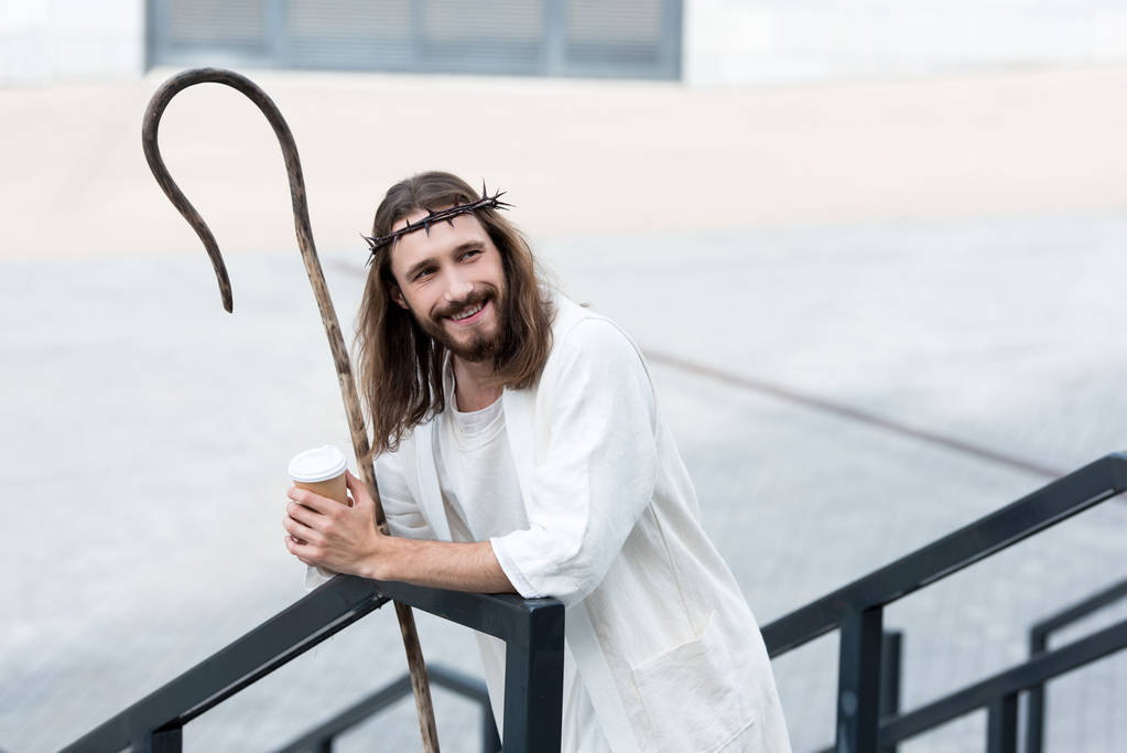 lächelnder Jesus in Robe und Dornenkrone mit Einweg-Kaffeetasse und an Geländer auf der Straße gelehnt - Foto, Bild