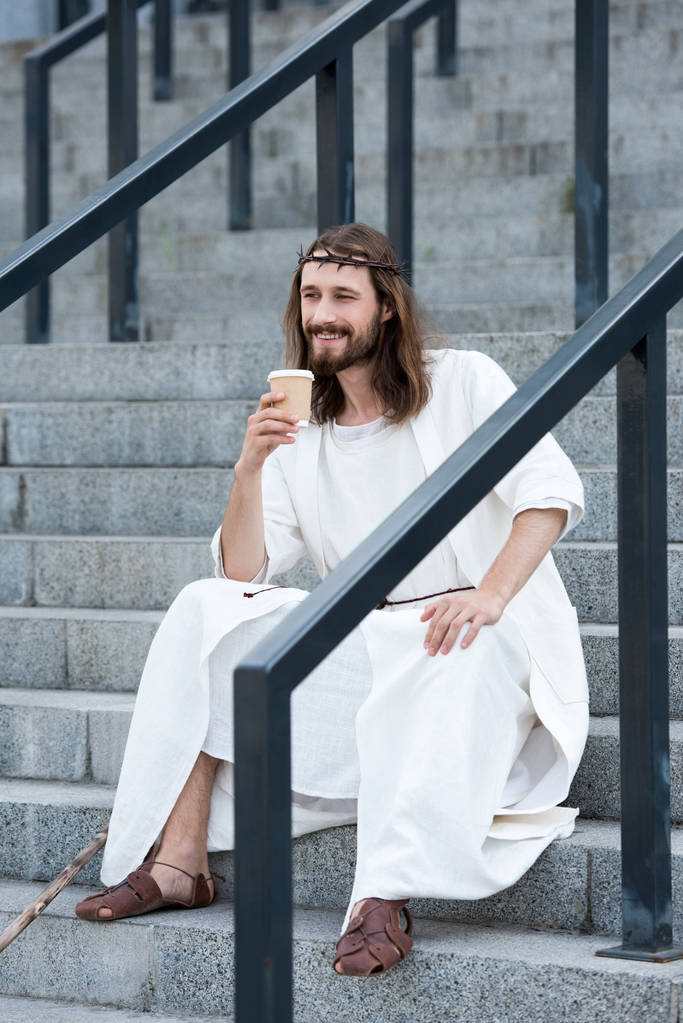 sorrindo Jesus em roupão e coroa de espinhos sentado em escadas e segurando copo de café descartável na rua
 - Foto, Imagem