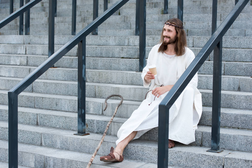 cheerful Jesus in robe and crown of thorns sitting on stairs and holding disposable coffee cup on street - Photo, Image