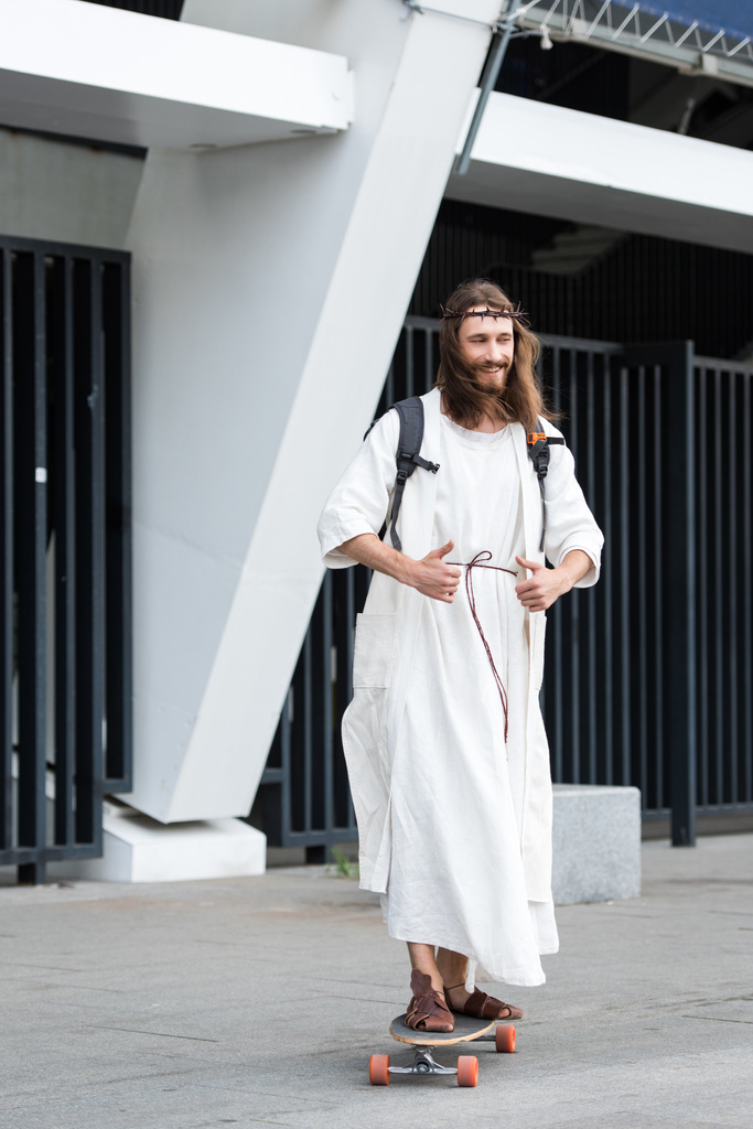 smiling Jesus in robe and crown of thorns skating on longboard on street and showing thumbs up - Photo, Image