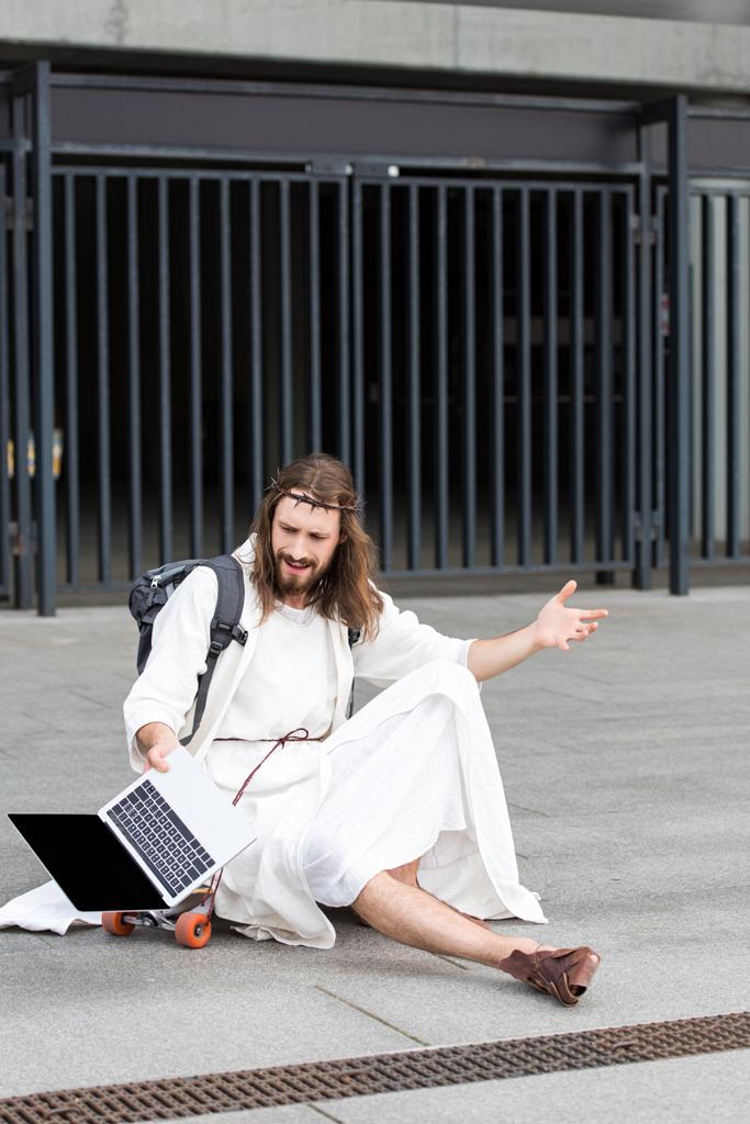 geïrriteerde Jezus in gewaad en kroon van doornen zittend op een skateboard en gebaren op laptop met leeg scherm in plaats - Foto, afbeelding
