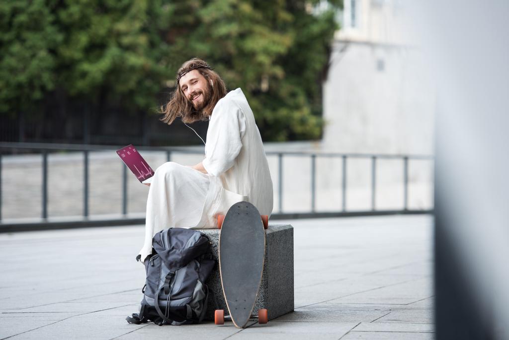 Jezus in de mantel en de kroon van doornen zittend op een steen en met behulp van laptop met medisch toestel op straat - Foto, afbeelding