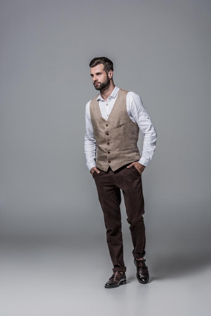 knappe bebaarde man poseren in trendy elegante vest op grijs - Foto, afbeelding