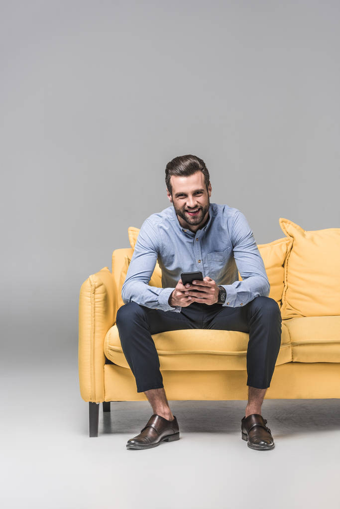 χαρούμενος άνθρωπος χρησιμοποιώντας smartphone και κάθεται στο κίτρινο καναπέ σε γκρι - Φωτογραφία, εικόνα