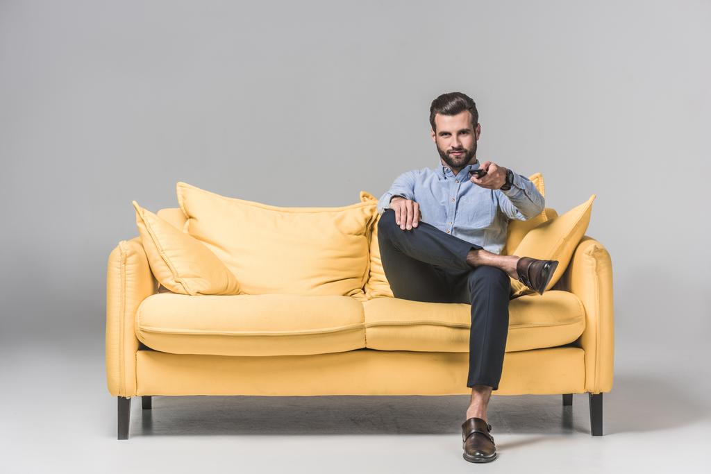 όμορφος χαμογελαστός άνθρωπος με τηλεχειριστήριο, βλέποντας τηλεόραση και κάθεται στον κίτρινο καναπέ σε γκρι - Φωτογραφία, εικόνα