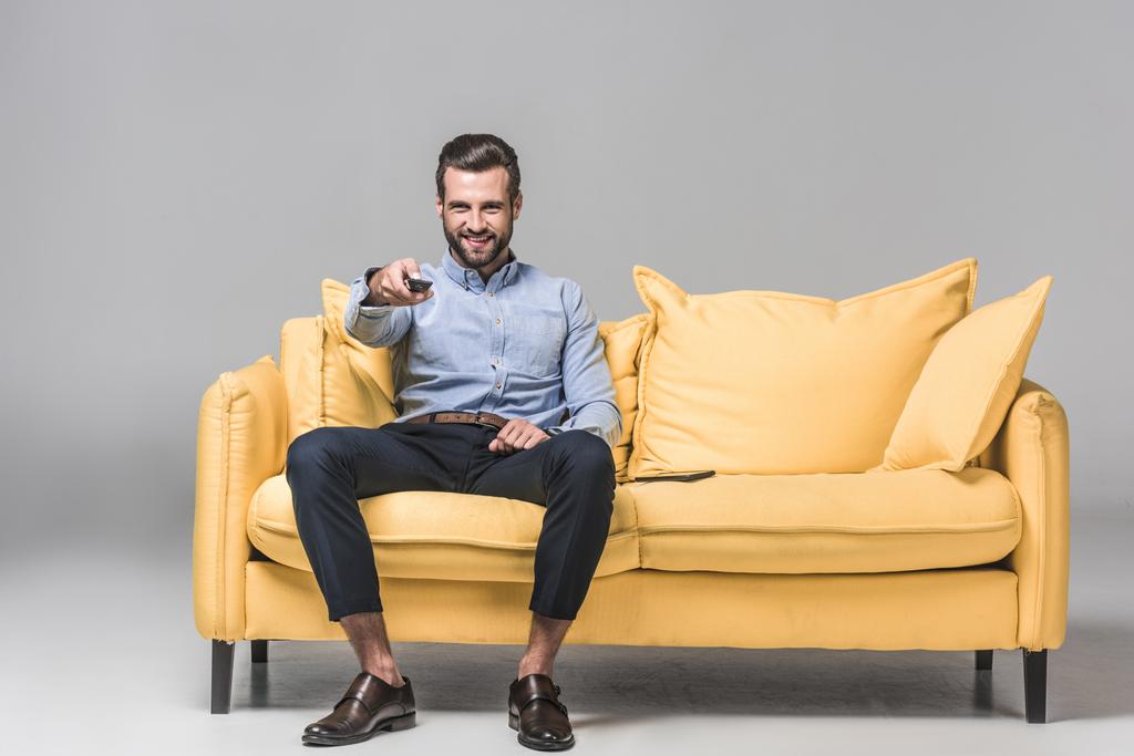 χαρούμενα επιχειρηματίας με τηλεχειριστήριο, βλέποντας τηλεόραση σε κίτρινο καναπέ σε γκρι - Φωτογραφία, εικόνα