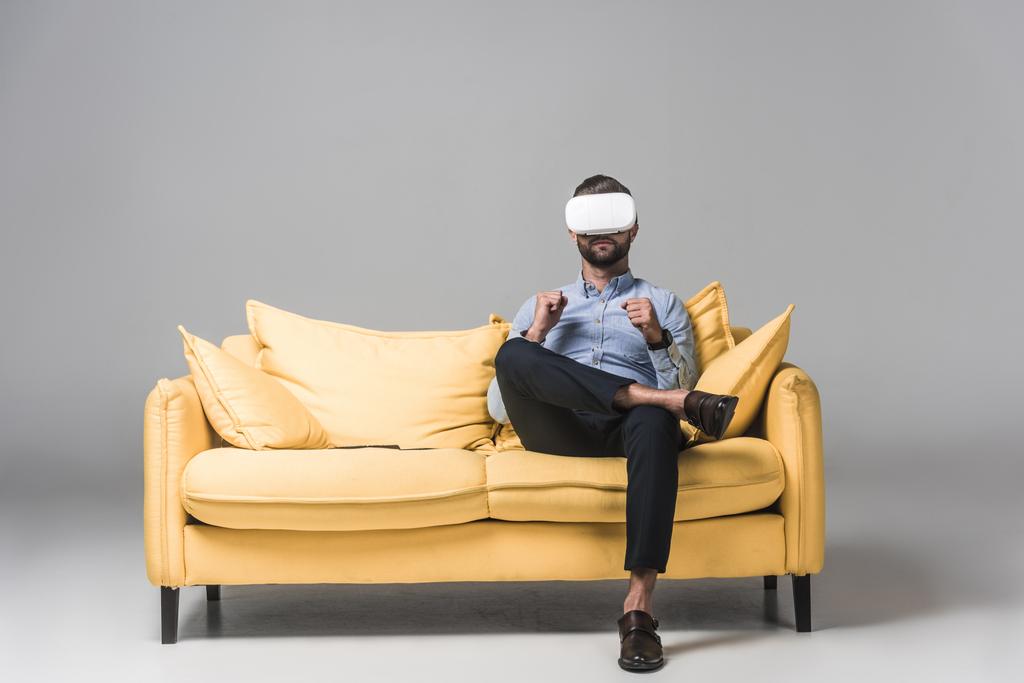 ο άνθρωπος χρησιμοποιώντας το σετ κεφαλής εικονικής πραγματικότητας και να κάθεται στο κίτρινο καναπέ σε γκρι - Φωτογραφία, εικόνα