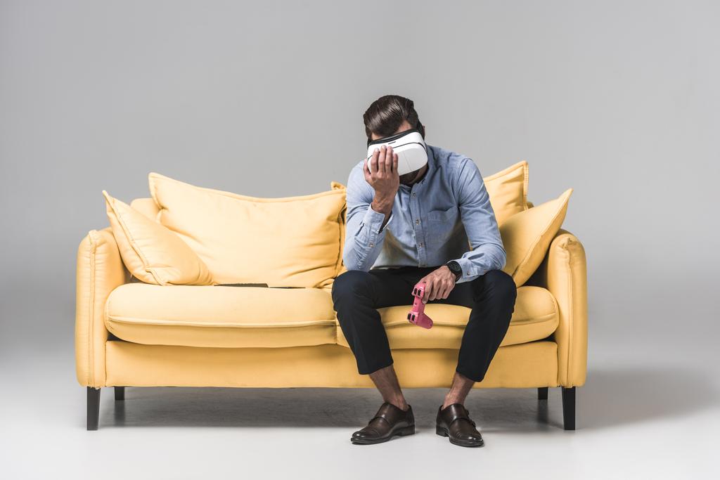 upset man lost in video game with joystick and virtual reality headset on yellow sofa on grey - Photo, Image