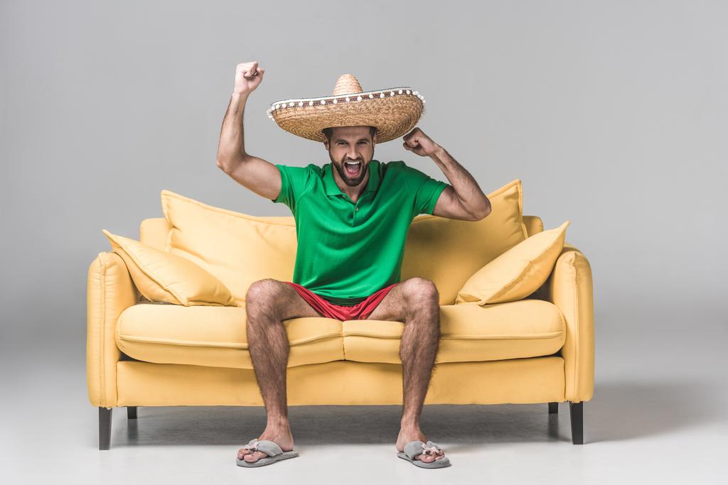 knappe man in Mexicaanse sombrero schreeuwen en vieren overwinning op gele sofa op grijs   - Foto, afbeelding