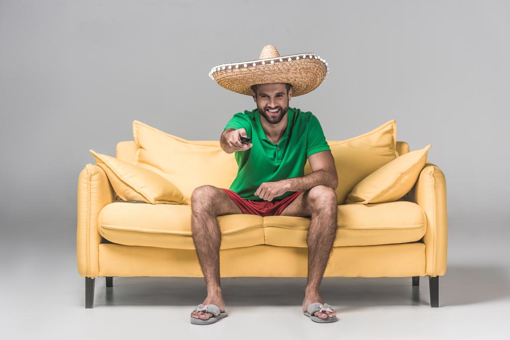 Lächelnder bärtiger Mann im mexikanischen Sombrero vor dem Fernseher mit Fernbedienung auf gelbem Sofa auf grau  - Foto, Bild