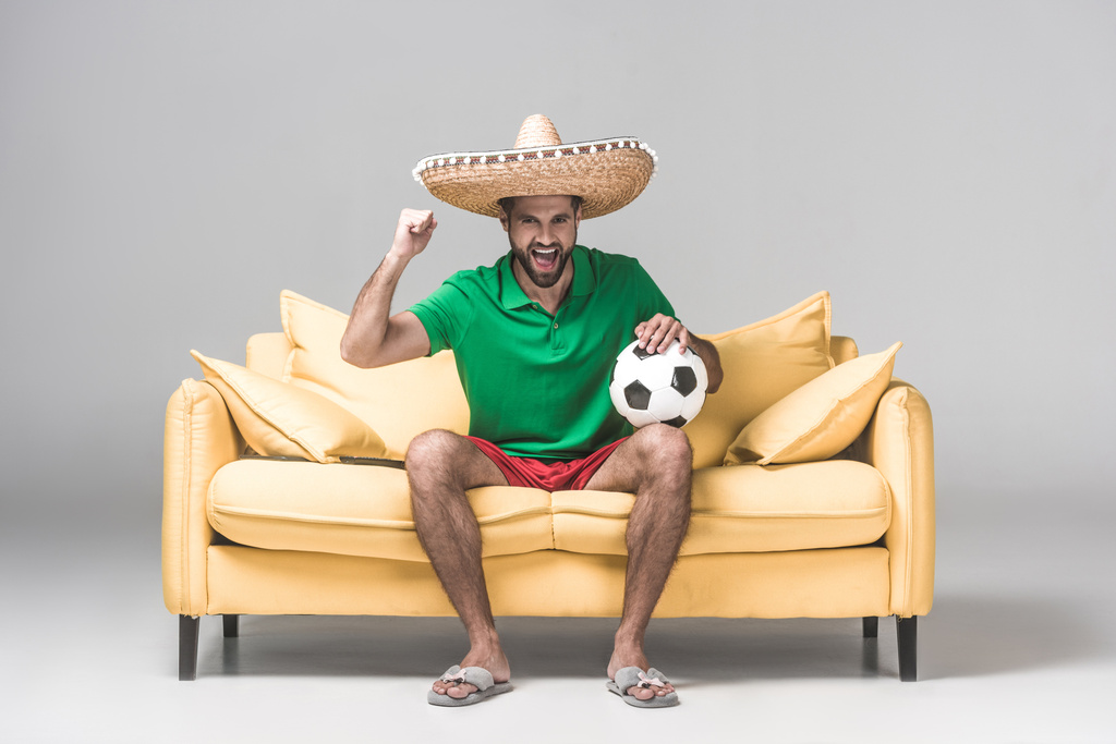 vidám ember, a mexikói sombrero néz foci mérkőzés labdát a szürke sárga kanapén ülve   - Fotó, kép