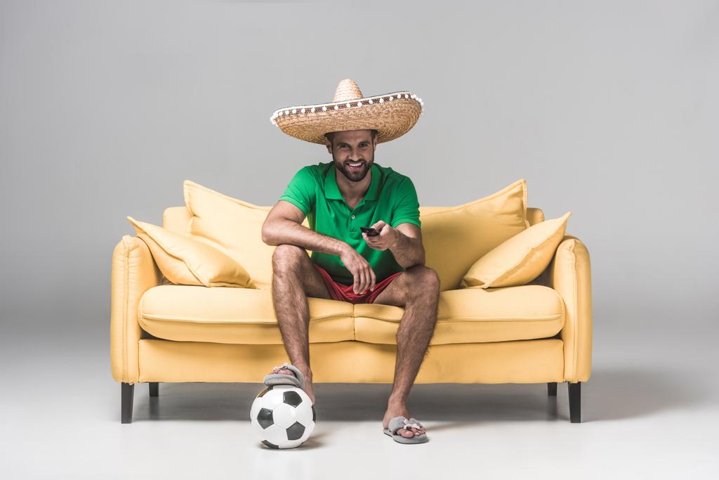 handsome mexican man in sombrero watching football match while sitting on yellow sofa with ball and remote control on grey   - Photo, Image