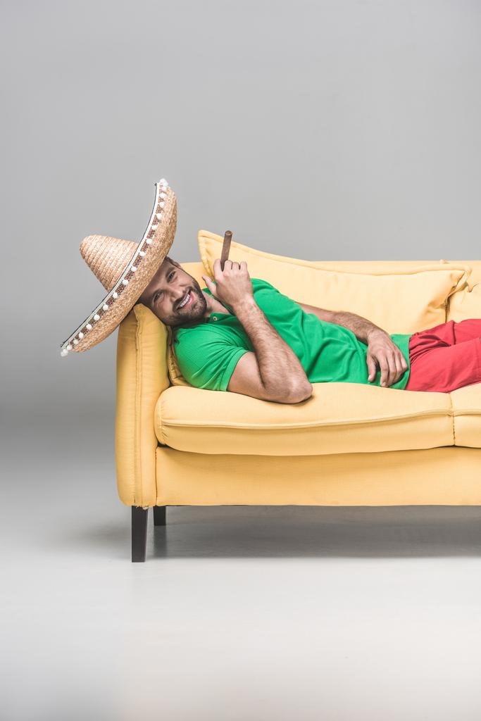 όμορφος άνδρας χαρούμενα στο μεξικάνικο σομπρέρο με πούρο που βρίσκεται στο κίτρινο καναπέ σε γκρι  - Φωτογραφία, εικόνα