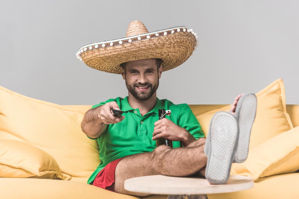 pohledný usmívající se muž v mexické sombrero sledování televize s dálkovým ovládáním, zatímco sedí na žluté pohovce s lahví piva na grey  - Fotografie, Obrázek